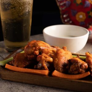 A plate of spicy buffalo wings served with carrot sticks and a dipping sauce, accompanied by a glass of beer, set on a dark table with a colorful napkin in the background at a cocktail bar.
