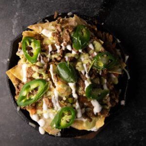 Top view of a skillet loaded with Mexican nachos topped with ground meat, melted cheese, jalapeno slices, and a drizzle of sour cream, shot on a dark background.