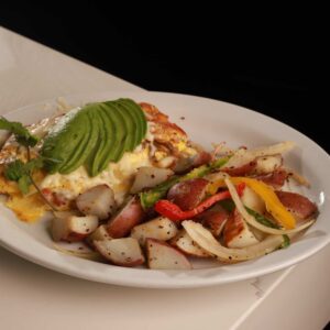 A plate of scrambled eggs topped with sliced avocado next to a side of roasted potatoes mixed with bell peppers, served on a white plate against a black background at a Mexican restaurant.