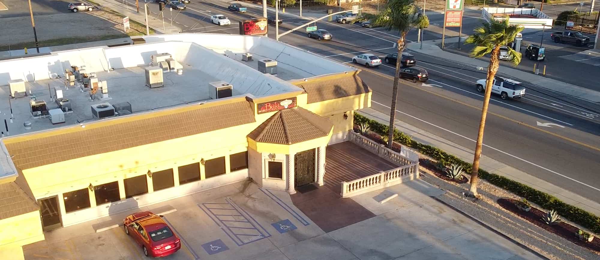 Aerial view of El Baja in Fontana, adjacent parking lot, and nearby road with moving vehicles. The setting sun casts long shadows across the scene.