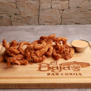 A wooden platter of crispy onion rings served with a side of dipping sauce, set against a stone wall background, with "el baja bar & grill" branded on the platter at the restaurant.