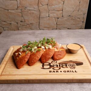Three golden-brown tacos filled with a variety of toppings, served on a wooden tray labeled "baja restaurant & grill", accompanied by a side of sauce, against a stone wall background.