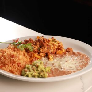 A plate of Mexican food featuring spicy chicken tinga tacos, red rice, refried beans, and guacamole, garnished with fresh cilantro, on a white plate against a black background.