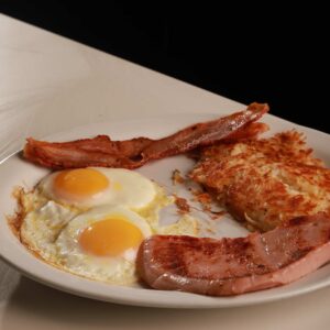 A traditional breakfast plate featuring two sunny-side-up eggs, crispy bacon strips, a grilled ham slice, and golden hash browns, served on a white plate at a Mexican restaurant.