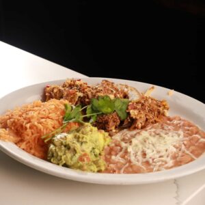 A plate of authentic Mexican food including rice, refried beans topped with cheese, and shredded meat garnished with a sprig of cilantro and a side of guacamole.