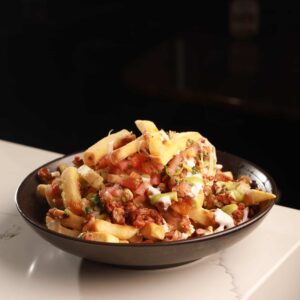 A bowl of loaded fries topped with ground meat, diced tomatoes, sliced pickles, and drizzled with a creamy sauce, served in a Mexican restaurant on a white surface against a dark background.