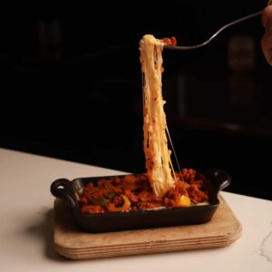 A person lifting a forkful of cheesy pasta from a black dish filled with baked ziti, revealing a stretch of melted cheese. The dish also contains pieces of meat and authentic Mexican peppers.