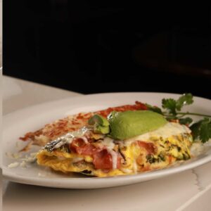 An omelette topped with slices of avocado and served with a side of hash browns on a white plate, garnished with fresh parsley, perfect for enjoying at our Mexican food-themed cocktail bar.