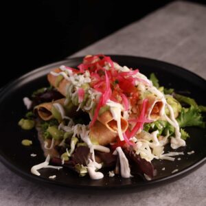 A plate with two crispy Mexican taquitos topped with shredded lettuce, cheese, pickled red onions, and salsa, served on a bed of mixed greens.