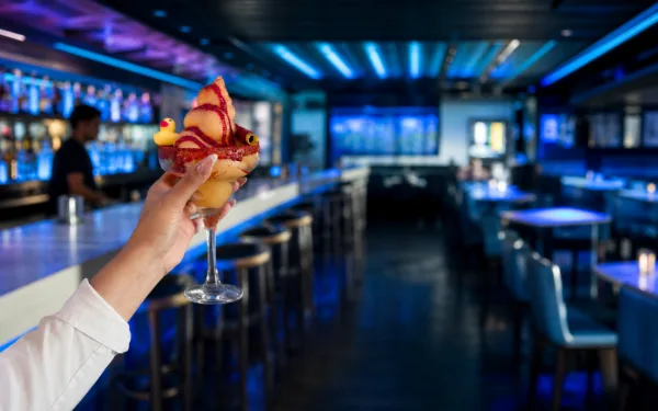 A hand holds a playful mango Soft Serve Margarita with a chili-coated rim, fruit garnish, and yellow duck inside El Baja.